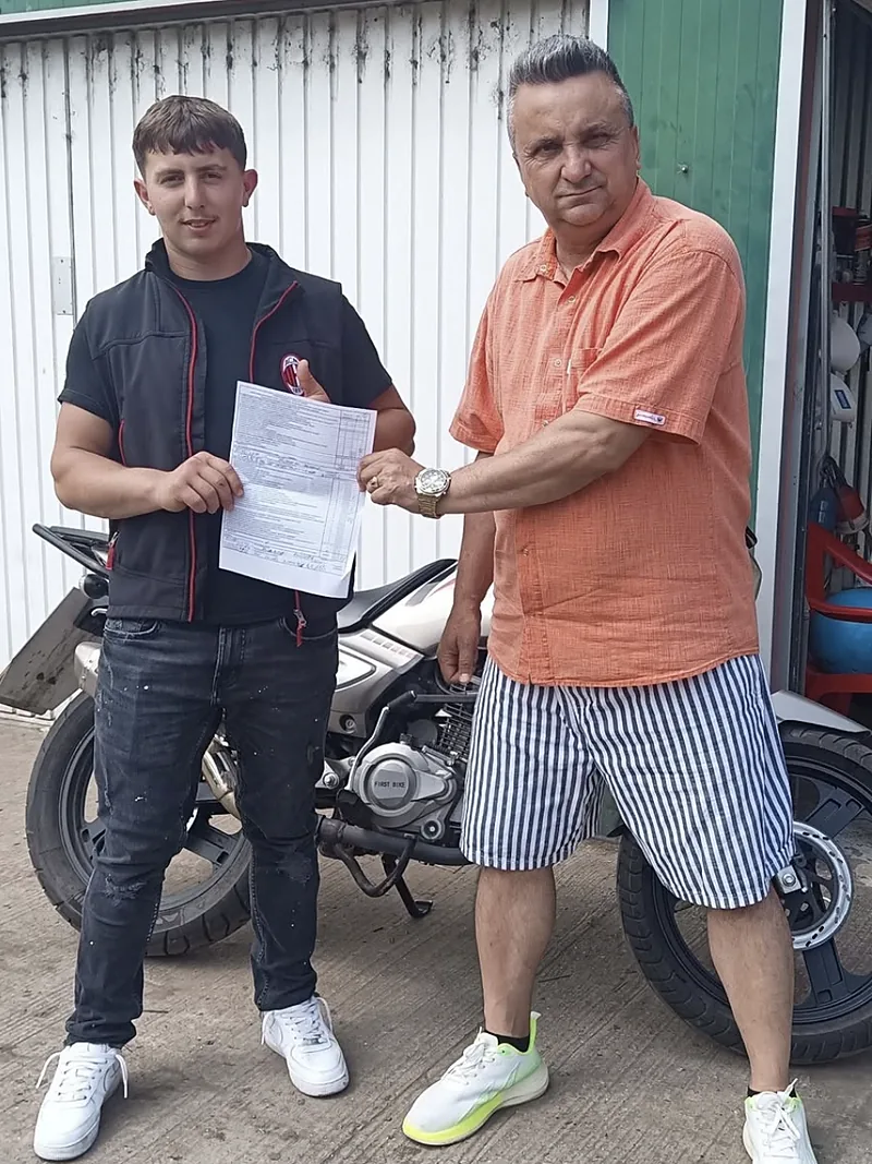 Cursant moto cu fișa de examen, alături de instructor, lângă motocicleta școlii